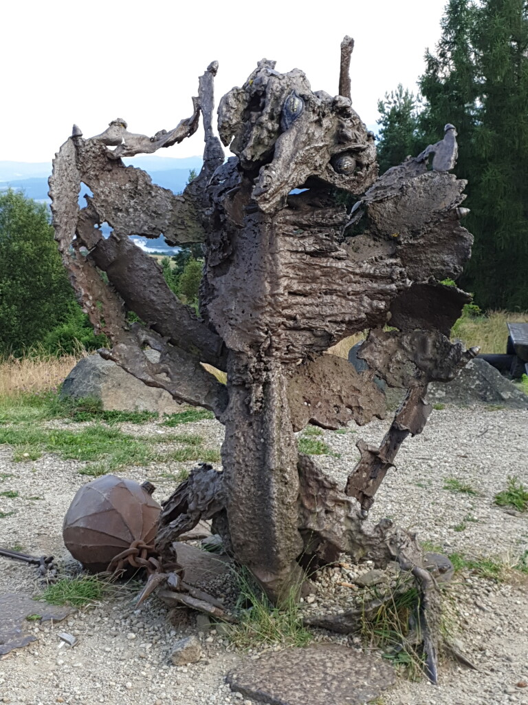 Photo of a dragon statue made of metal, looking rusty and torn. There are visible eyes made of small metal spheres and big metal ball attached to its leg.