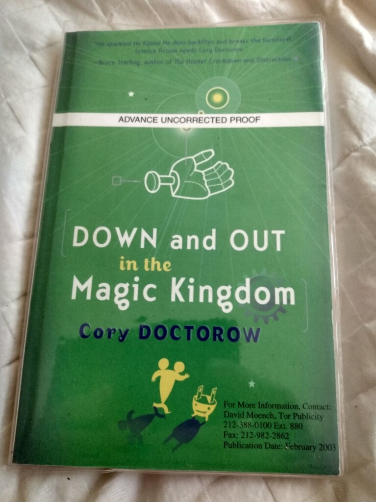 A green book cover is shown laying on an off white comforter, it is Down and Out In The Magic Kingdom by Cory Doctorow.