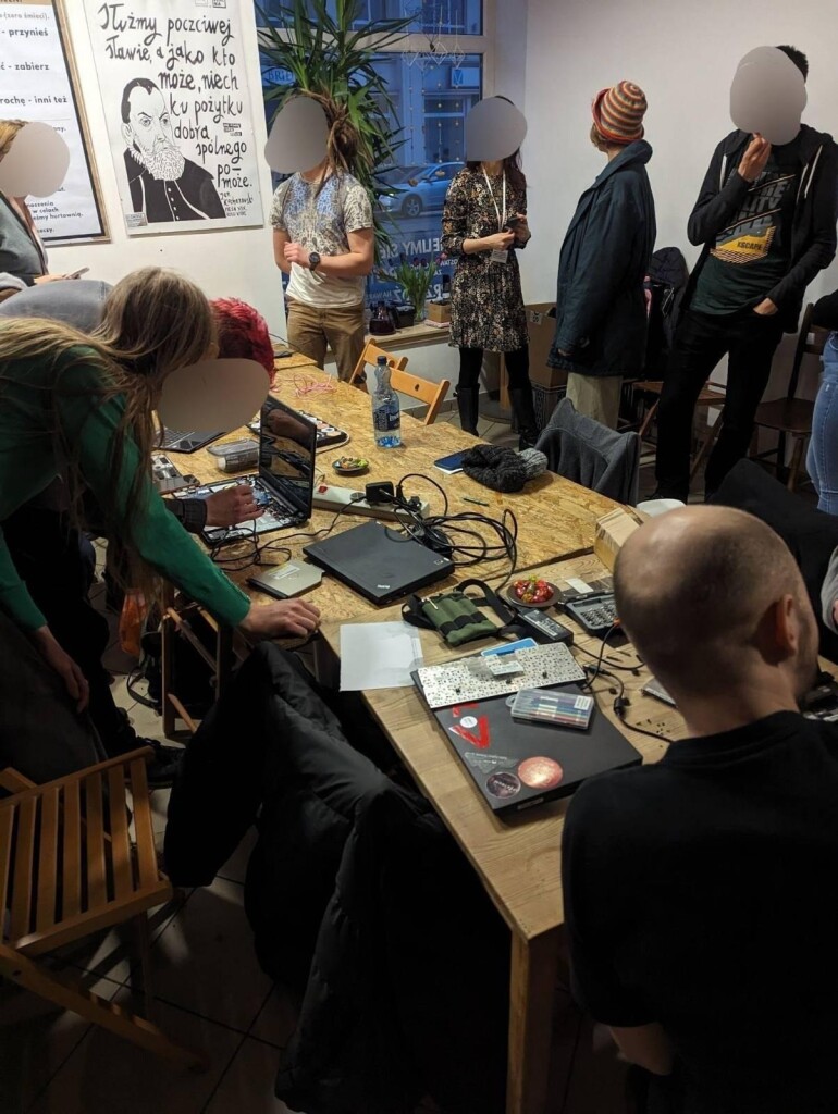 Pokój w Po:dzielni. Grupa ludzi znajduje się przy stołach pełnych laptopów i innej elektroniki. Hakerzy pomagają naprawić sprzęty przyniesione przez mieszkańców. 