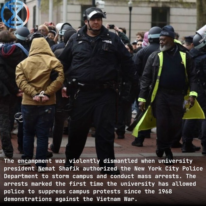 A police officer stands in the foreground. Behind them is someone with their back to the camera, with hands cuffed behind their back, part of a crowd with other people and police officers. The police are wearing helmets implying they may face violence, but the crowd is peaceful.