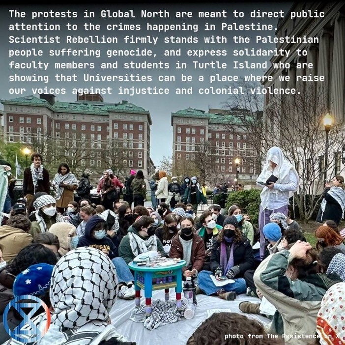 Photo from The Resistance. The gathering on the Colombia lawn. Many are seated on a ground sheet, around a space with a table in the centre on which there is food. Most of those visible are wearing headscarves and/or keffiyeh scarves. Many are wearing face masks (surgical masks or N95/FFP2 masks).
