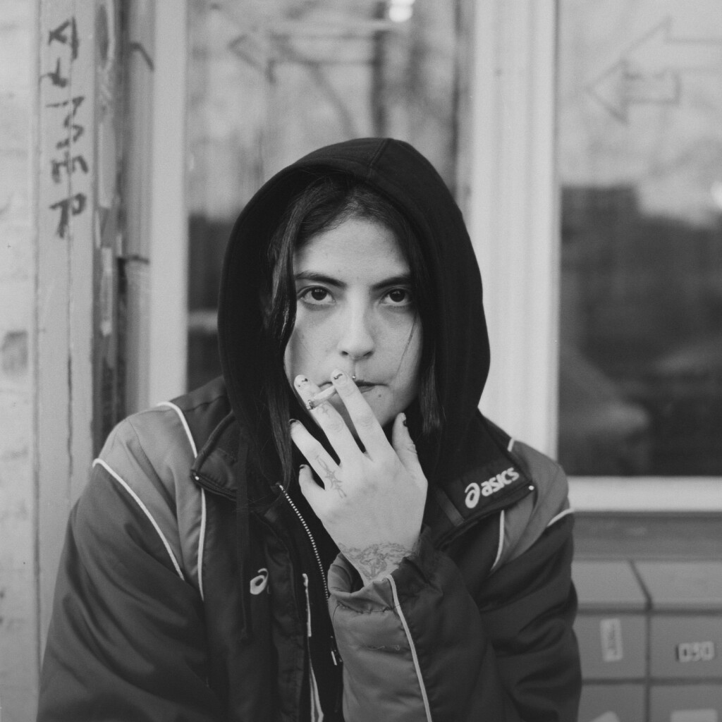 b/w street portrait of a young person with light skin, wearing a hoodie and smoking a cigarette. their irises are very dark, almost entirely black