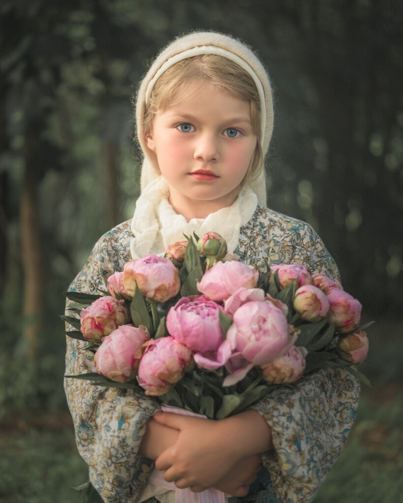 A child wearing a cream headscarf and a green floral shirt. Holding a bouquet of pink peonies. 