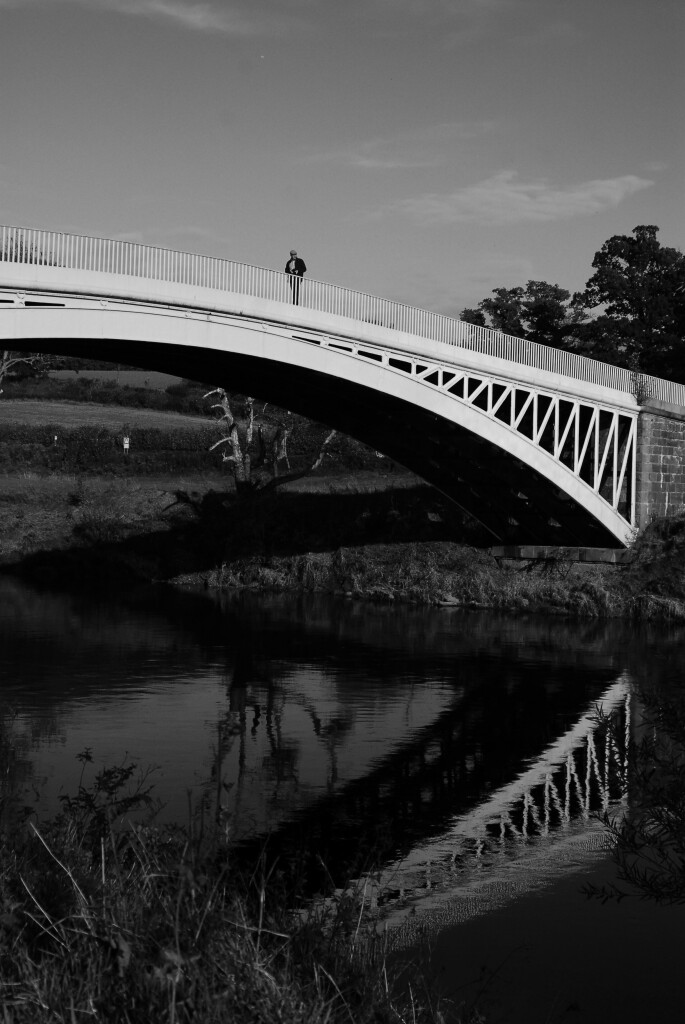 A black and white photograph of a victorian age iron bridge near Bigsweir which straddles the border between England and Wales over the river Wye. It's reflected in the calm water it spans, and a man looks over the side. 