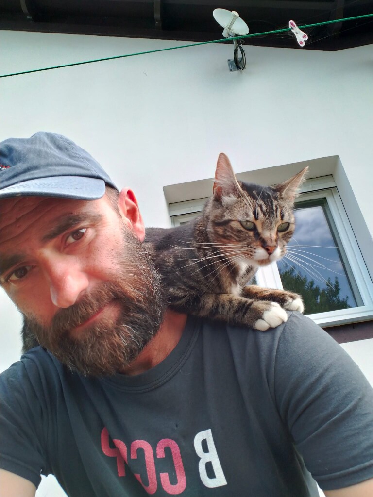 Selfie: it's me wearing a blue cap and a black t-shirt with Lilith, a tabby small cat, on my shoulders. I'm staring at the camera and her to the right side of the pic