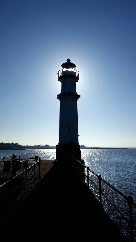 A lighthouse at the end of a pier, silhouetted by the sun behind it. The sky is a deep cloudless blue, and the sea a similar blue with gentle ripples.