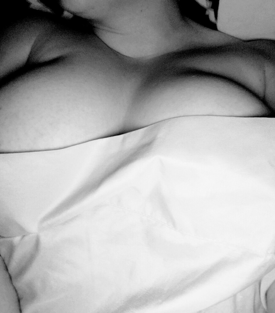 A black and white photo of a woman laying naked in a bed. She's covered by a sheet up to her breasts. You can see the shape and soft curves of the breasts but no nipples 
