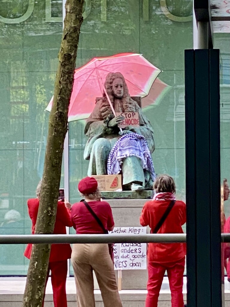 Statue of 18th century judge, but now with umbrella in colors of a water melon, and a sign “stop genocide”