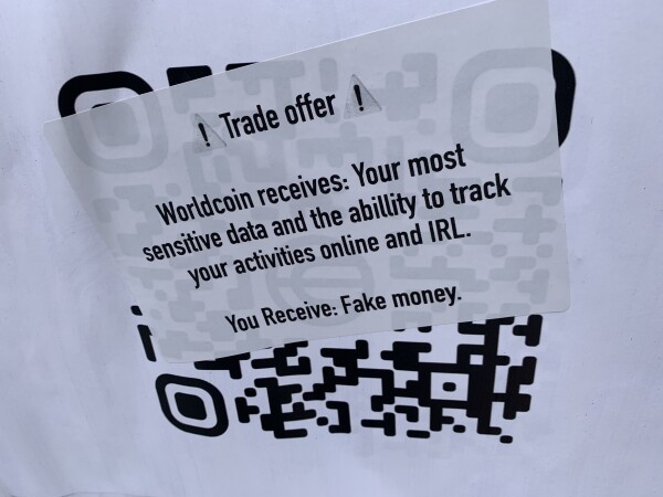 A sticker placed over the QR code on the Worldcoin flyer. The sticker says “Trade offer. 
Worldcoin receives: your most sensitive data and the ability to track your activities online and IRL. 
You receive: Fake money”