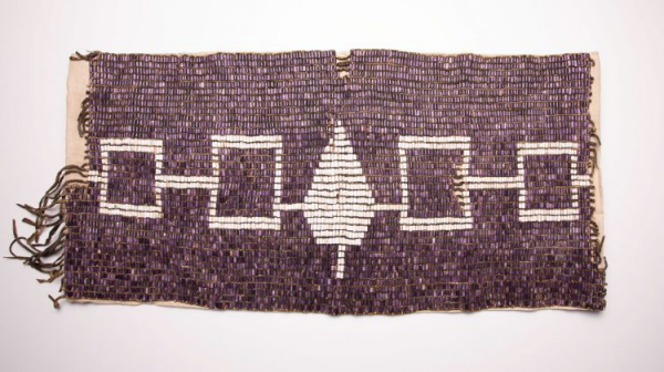 A photograph of the Hiawatha Belt, the emblem of the Haudenosaunee or Iroquois confederacy. It is a belt made from wampum beads, densely woven together. It depicts a line of four interconnected rectangles centered on a single stylized tree, all in white against a rich purple background.