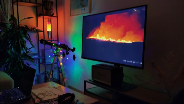Photo of a cozy corner of an apartment, showing an orange lava lamp, a telescope with some Christmas lights on it, and a flat screen TV with a stream of the eruption on.