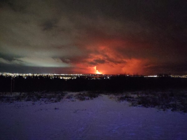Night (and grainy) photo of a snowy hill, some dark bushes, city lights, and a distant volcanic eruption, lighting up the sky blood-red.