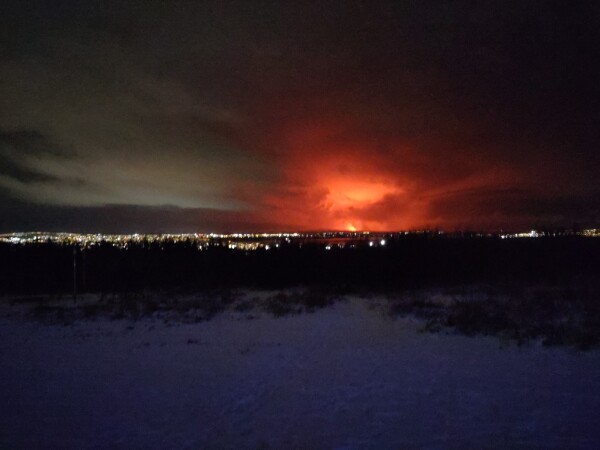 Night (and grainy) photo of a snowy hill, some dark bushes, city lights, and a distant volcanic eruption looking definitely menacing. Dark clouds giving a strong contrast to the orange-red eruption.