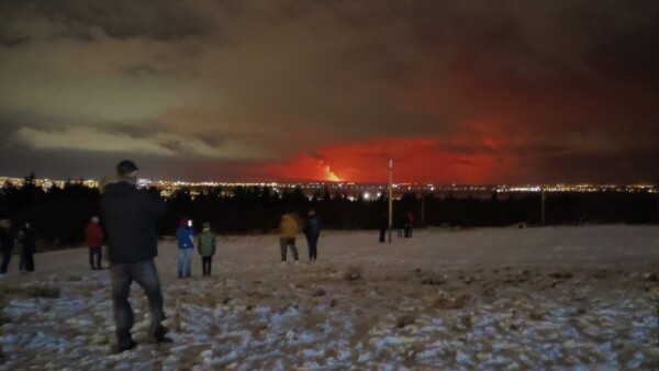 A bunch of people gathered on a snowy hill overlooking a city. Distant eruption visible.