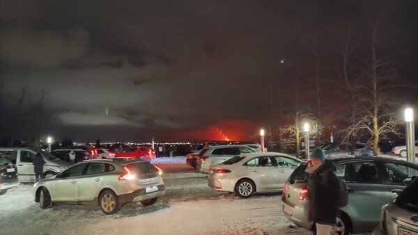 Snowy, well-lit car parking lot with a bunch of cars manoeuvring. Volcanic eruption visible in the distance.