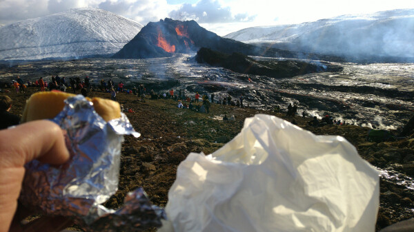 Erupting volcano in the background, with lava field visible and a few dozen people watching.

Foreground: a hand holding a partially tinfoil-wrapped sandwich, a bit blurry (focus on the volcano).

Snowy hills behind the volcano cone.