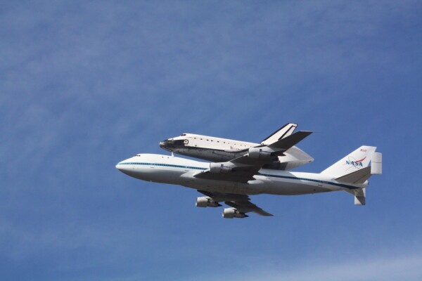 A picture of the Space Shuttle Endeavour, atop the carrier 747 aircraft, flying through a nearly cloudless blue sky.
