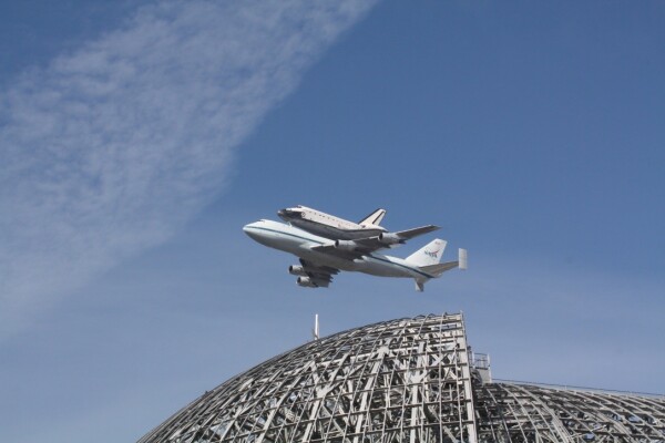 A picture of the Space Shuttle Endeavour, atop the carrier 747 aircraft, flying through a nearly cloudless blue sky. In the lower foreground is a picture of the massive blimp hangar superstructure at Nasa / AMES.
