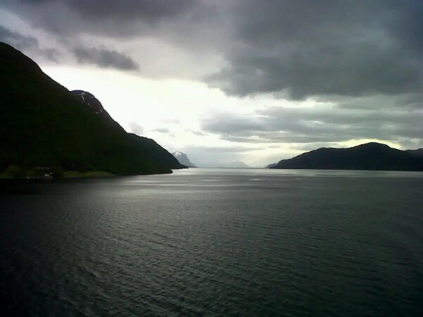 Moody shot of a Norwegian fjord. Mountains roll down to flat water, oppressive clouds fill the sky. The shot isn’t desaturated, but looks like it is. 