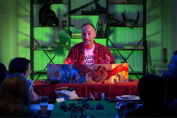 A colorful and dramatic photograph of a man running a game of Dungeons & Dragons, emoting and gesturing with players in the foreground.