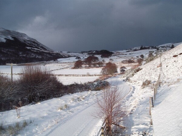 View down a snow-covered rural valley with a shallow slope on the left and a less snow covered wooded slope on the right. A snow covered road winds along the bottom of the slope to the right with fields to its left across the floor of the valley. There's bright sunshine from left with a very ominous looking dark snow shower approaching from the north (up the valley).