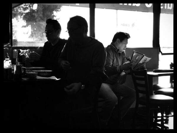 A black and white photo of 3 men sitting in a Vietnamese restaurant. Two men are facing the left and are silhouetted in the foreground eating their food. The other man is behind them and to the right, lit in the morning sun as he reads a menu.