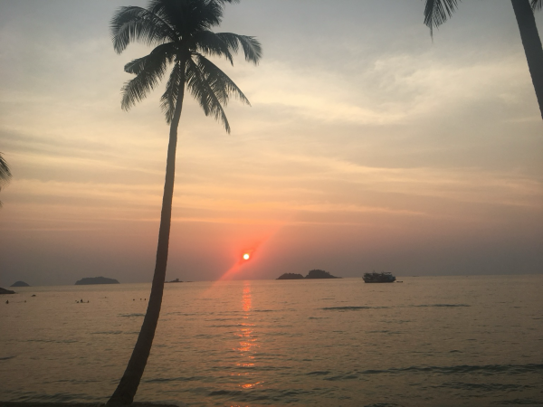 The orange sun close to the horizon over a calm sea. There are some small islands in the distance and a coconut palm in the foreground