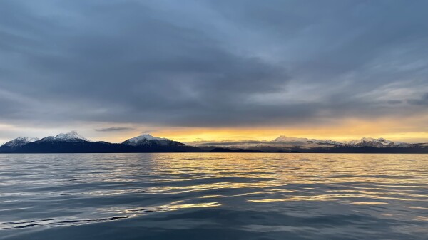 A bright yellow sunset, sandwiched between a dark grey-blue cloudy sky and a dark blue sea with gentle waves. There are snow-capped mountains on the horizon.