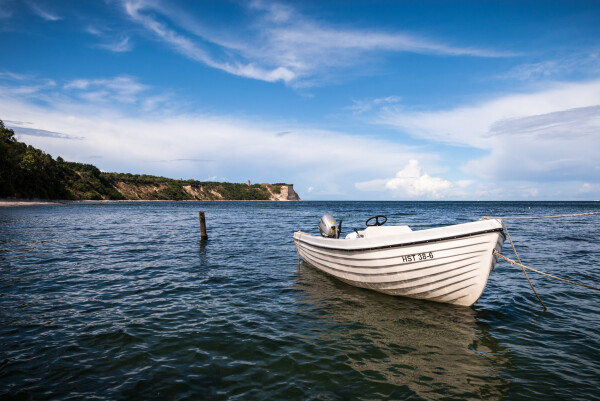 a small white fishing boat sitting in the water in front of a very high coastline (steilküste rügen, just east of Arkona for those who know). the sky is very blue with only some clouds. the blue color is reflected in the otherwise dark water.