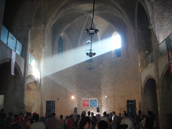 DJ playing to a crowd in a former church, with the sun shining through one of the windows, reflecting on the wall to light up the DJ.
