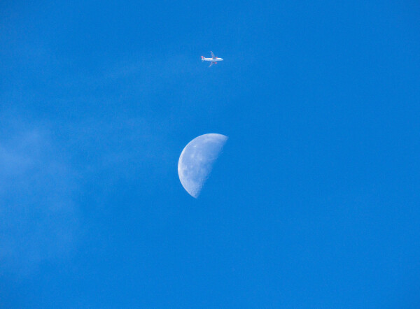 A perfectly blue sky with a half moon dead centre and directly above it, a passenger jet flying towards the right of the image. 