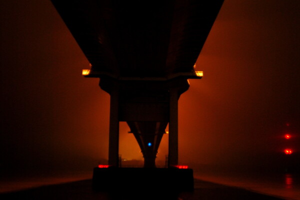 The underside of the Severn Bridge crossing in South Wales on a foggy night. It carves out an ominous shadow against bright orange lighting as the fog is lit by the lights of the bridge above