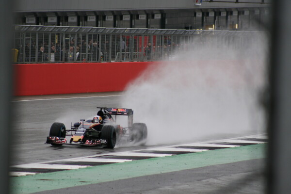 A Formula 1 car (Toro Rosso) shot through a gap in a fence, with spray from the rain blowing up behind it