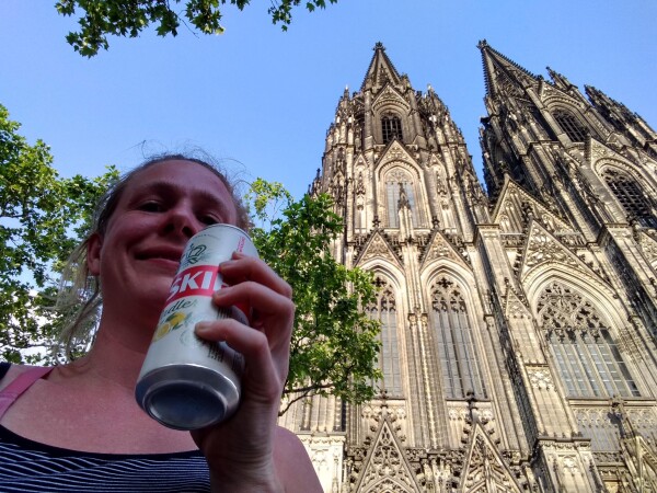 Selfie holding a can of radler, smiling, in front of the Kölner Dom. It's sunny and the Dom is lit, whereas I'm in the shadows. Some trees.