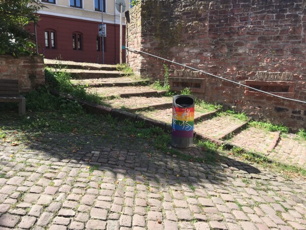 Rainbow trashcan in between old cobblestone and red sandstone buildings.