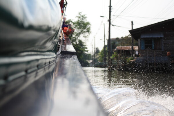 A view of a canal in Bangkok, taken with the camera resting on the outside frame of a long-tail boat moving at speed along the canal. The shot is predominantly the right hand side of the boat, and a large spray of water, with some dwellings on the side of the canal.