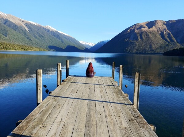 A small person sits at the end of a wooden pier extending into a lake with mountains on either side