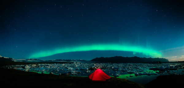 The image captures a stunning night scene featuring the Northern Lights (Aurora Borealis) over a landscape filled with ice and water. In the foreground, a red tent glows warmly, providing a stark contrast against the cool colors of the surroundings. The tent is pitched on a grassy patch near the edge of an icy lake or fjord, dotted with chunks of ice floating on the water's surface.

The sky is clear and filled with stars, creating a magnificent backdrop for the vibrant green and blue hues of the Northern Lights that stretch across the horizon. The light from the Aurora illuminates the landscape below, highlighting the rugged mountains in the distance and the icy terrain. The overall scene is serene and otherworldly, combining the raw beauty of nature with a sense of adventure and tranquility.
