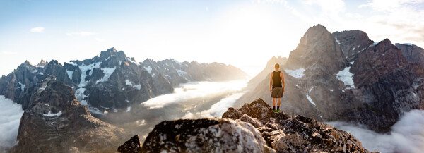 The image shows a breathtaking mountain landscape during what appears to be sunrise or sunset. A lone figure, dressed in a black tank top and gray shorts, stands on a rocky outcrop, gazing out at the scene before them. The person is positioned in the right foreground, facing away from the camera, which captures the expanse of rugged, jagged peaks in the background. The mountains are partially covered with patches of snow and glaciers, and the valleys below are filled with a layer of clouds, giving a sense of immense height and vastness. The sky is clear with a few scattered clouds, and the sunlight creates a warm glow on the peaks and the figure, adding to the majestic and serene atmosphere of the scene.