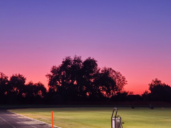 A sunset after football practice at the college I used to work at. The sky is varying shades of hot pink through soft violet, the grass is a vivid bright green. The trees lining the river bank look black against the bright sky. In the foreground there is a metal water pumper 