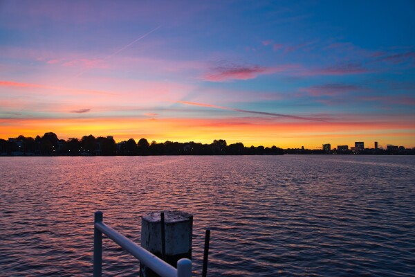 view of the Alster in Hamburg. Early morning sunrise, the sky is purple, blue and orange. the skyline is visible on the horizon, water in the lower half, a piece of a wooden pier in the bottom.