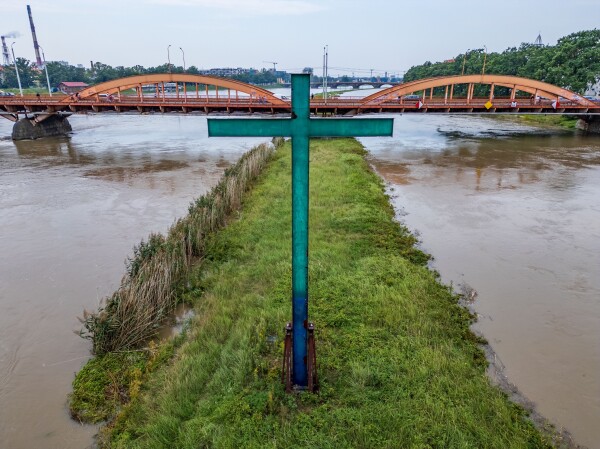 A large green cross stands prominently in the foreground, surrounded by tall grass and reeds. In the background, an arched bridge spans over a river, with pedestrians visible on it. The river's water is murky, and the scene is set under