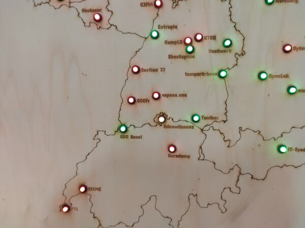 A detail view of a map laser-engraved into plywood, showing the area of Switzerland, south-western Germany and northeastern France. In the map there are multiple circular LEDs, each in a small black socket, glowing either green or red. Next to each LED there is the name of a hackerspace engraved into the wood. The colors of the LEDs indicate whether the corresponding LED is currently open or closed.