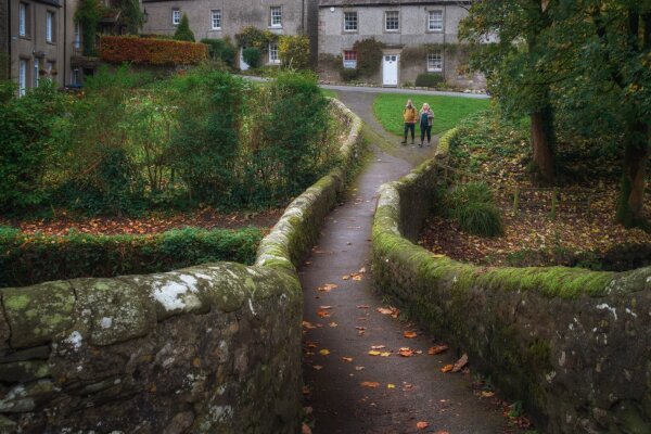 A moss-covered stone bridge with curved walls leads to traditional Yorkshire stone cottages in the village of Clapham, whilst two walkers pause on the path ahead. The narrow footbridge, dusted with autumn leaves, curves gently upward between weathered walls topped with verdant moss, creating a charming scene typical of Yorkshire Dales architecture, with the grey-stone houses and their distinctive sash windows providing a quintessentially British backdrop.