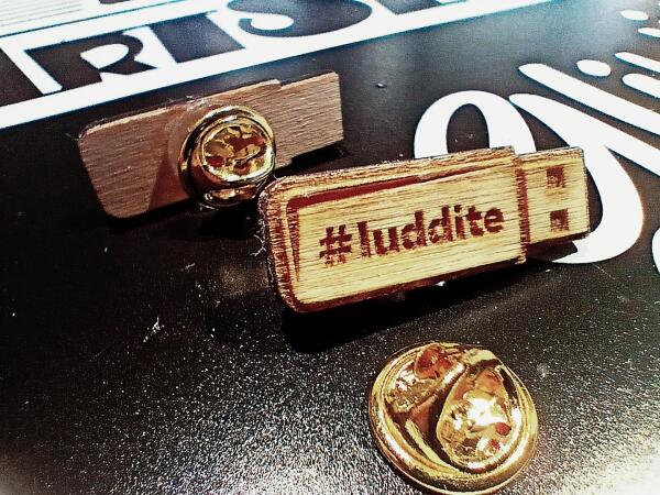 Małe przypinki, wykonane z grawerowanej laserem sklejki, w kształcie pendrive z napisem #luddite Leżą na podkładce z logo firmy wArtSztat.