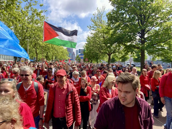 Protestors dressed in red with a palestinian flag, in the sun, in between britht green trees